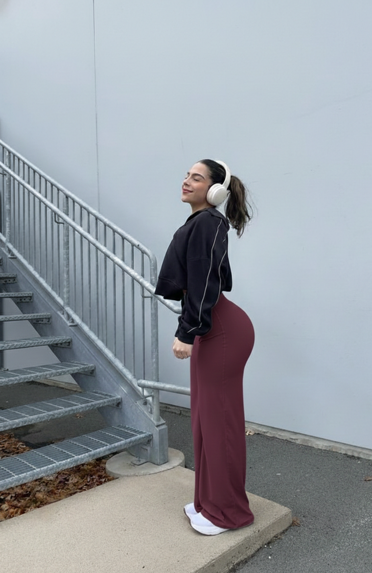 Person wearing a black sweatshirt and maroon pants standing on a sidewalk with a light blue wall and metal railing in the background.